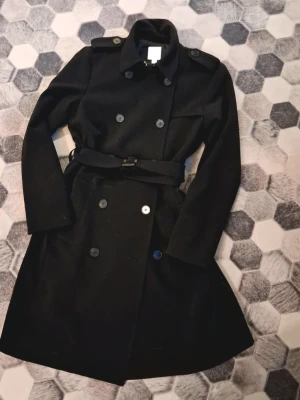 Svart trenchcoat från H&M med bälte - Klassisk svart trenchcoat från H&M i storlek 32. Jackan har dubbelknäppning, axelklaffar och ett matchande bälte i midjan. Långärmad modell med krage och knappar framtill. Perfekt för dig som gillar stilrena och tidlösa plagg.