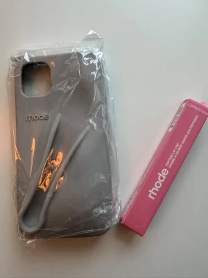 Grått Rhode mobilskal med rosa läppglans  - Snyggt grått mobilskal från Rhode med läppglans för IPhone 11. Oöppnat i kartong. Köpte fel storlek så säljer vidare. 