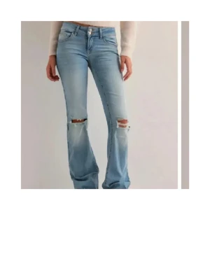 Ljusblå bootcut jeans med slitningar - Säljer ett par ljusblå bootcut jeans med slitningar över knäna och klassisk femficksdesign. Jeansen har låg midja och är tillverkade i denim med lite stretch för skön passform. Perfekta för en avslappnad och trendig look. Från Nelly, lite trasiga i slutet men inget som syns mycket!