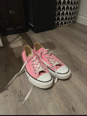 Rosa Converse sneakers låga - Säljer ett par klassiska Converse sneakers i rosa med vita snören och tåhätta. Skorna har låg profil och rund tå, samt den ikoniska vita sulan med svart rand. Tillverkade i slitstark bomullscanvas och passar perfekt till en avslappnad stil.