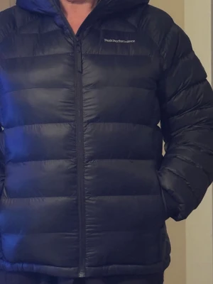 Svart dunjacka Peak Performance - Snygg svart dunjacka från Peak Performance med huva och dragkedja framtill. Jackan har en quiltad design och är perfekt för kalla dagar. Klassisk pufferstil med lång ärm och diskret logga på bröstet. Passar dig som vill ha både stil och värme.