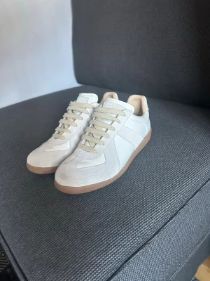 Maison Margiela skor  - Stilrena beige sneakers med ovandel i mocka och skinn, samt ljusbruna gummisulor. Klassisk låg modell med snörning och rund tå. Perfekta för en clean och avslappnad look.
