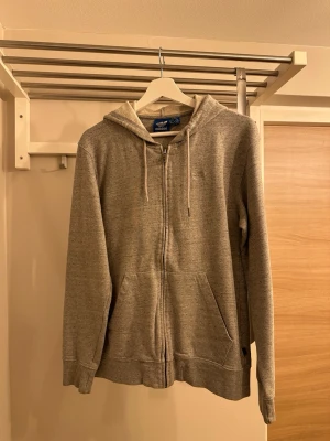 Grå zip hoodie från Adidas - Säljer en stilren grå zip hoodie från Adidas med dragsko i huvan och två fickor framtill. Klassisk Adidas-logga på bröstet och mjukt material som känns skönt mot huden. Perfekt för en avslappnad look och enkel att matcha med andra plagg.