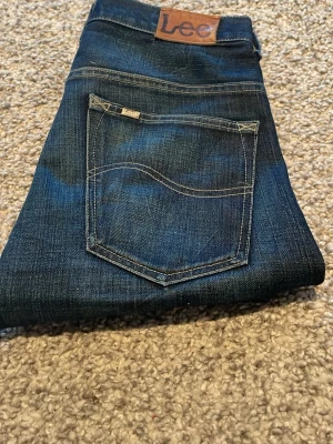 Mörkblå Lee jeans med raka ben - Säljer ett par klassiska mörkblå jeans från Lee med raka ben och snygga kontrastsömmar. Jeansen har två bakfickor och en Lee-lapp i läder baktill i midjan. Perfekta till en avslappnad stil och tillverkade i slitstarkt denimtyg.