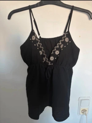 Topp ifrån shein - Snygg svart linnetopp med tunna axelband och vit brodyr i blommönster längs v-ringningen. Toppen har en liten knytning framtill och är perfekt för varma dagar. Passar till både jeans och kjol för en avslappnad look. Aldrig använd fint skick dock är lapparna avklippta