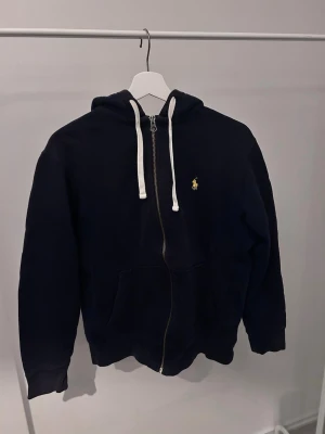Marinblå hoodie från Polo Ralph Lauren - Snygg marinblå hoodie med dragkedja från Polo Ralph Lauren. Klassisk design med vit dragsko i huvan, fickor framtill och den ikoniska gula loggan broderad på bröstet. Perfekt för en avslappnad och stilren look.