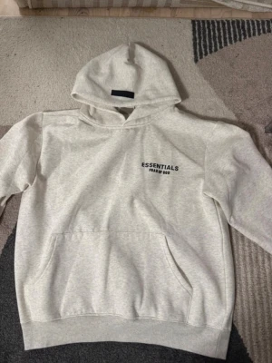 Grå Essentials Fear of God hoodie - Snygg ljusgrå hoodie från Essentials Fear of God med svart logga på bröstet och ryggen. Klassisk känguruficka framtill, ribbade muddar och stor huva. Perfekt för en avslappnad streetwear-look. Mjuk bomullsblandning för extra komfort.