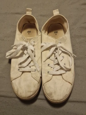 Vita sneakers från H&M med snörning - Klassiska vita sneakers från H&M i storlek 41. Skorna har rund tå, platt sula och snörning med breda vita skosnören. Tillverkade i bomullstyg med metallöljetter för snörena. Perfekta för en clean och enkel stil.