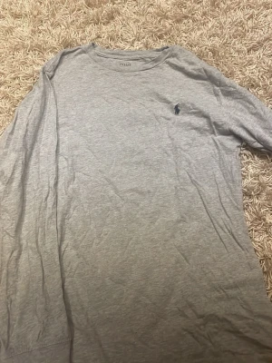 Grå långärmad tröja från Polo Ralph Lauren - Snygg grå långärmad tröja från Polo Ralph Lauren med klassisk rund halsringning och den ikoniska mörkblå logotypen broderad på bröstet. Tillverkad i mjukt bomullsmaterial som känns skönt mot huden. Perfekt för en avslappnad och stilren look.