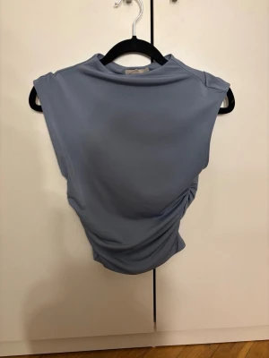 Ljusblå ärmlös topp med drapering - Trendig ljusblå topp med draperad detalj på sidan. Toppen är ärmlös och har en mjuk, stretchig känsla som ger en snygg passform. Perfekt för att skapa en stilren look med en twist.