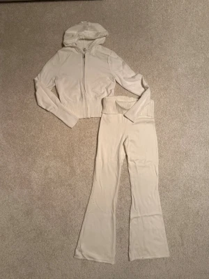 Vit loungewear-set från H&M - Mysigt loungewear-set från H&M i vitt. Setet består av en zip-hoodie med ribbade muddar och huva samt matchande byxor med bred ribbad midja och utsvängda ben. Perfekt för chill dagar hemma. Materialet är mjukt och stretchigt för extra komfort. Mycket fint skick, knappt använt.