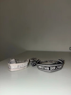 Christian Dior J'Adior armband - Två snygga Christian Dior J'Adior armband i tyg, ett i svart med vit text och ett i beige med vit text. Båda har tofsar och justerbar knytning. Perfekta för att ge din outfit en lyxig touch och visa din stilkänsla. Helt oanvända. 200kr styck 350kr för båda.