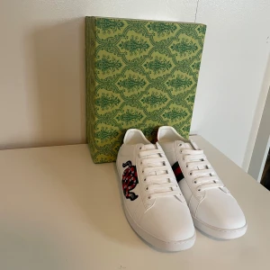 Vita Gucci sneakers med orm-motiv - Snygga vita sneakers från Gucci med ikoniskt orm-motiv i rött, svart och grönt på sidan. Skorna har röda och gröna metallic-detaljer på hälen och klassisk snörning framtill. Tillverkade i skinn för en lyxig känsla och stilren look. Tveka inte på att höra av dig om du har några frågor eller liknande 😊