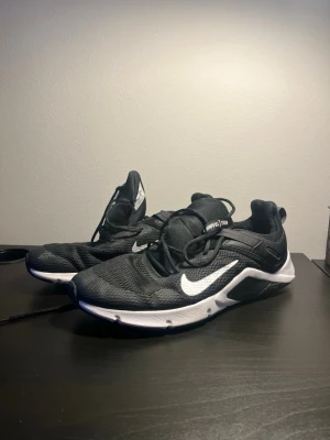 Svarta Nike skor  - Snygga svarta Nike sneakers med vit sula och klassisk vit swoosh på sidan. Ovandelen är i mesh för bra andningsförmåga och skon har snörning. Perfekta för en sportig och modern stil.