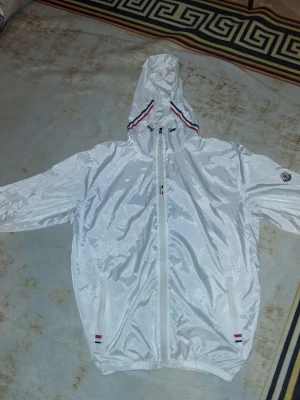 Moncler vindjacka med huva - Priset kan diskuteras. Snygg vit vindjacka från Moncler med huva och hel dragkedja framtill. Jackan har Moncler-logga på ärmen, två fickor med dragkedja och detaljer i rött, vitt och svart vid huva och fickor. Perfekt för blåsiga dagar och enkel att matcha. Har kanppast använt. 