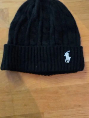 Svart mössa från Polo Ralph Lauren - Snygg svart ribbstickad mössa från Polo Ralph Lauren med klassisk vit broderad logga framtill. Mössan har vikt kant och är tillverkad i mjukt material som håller dig varm under kalla dagar. Perfekt accessoar för en stilren look.