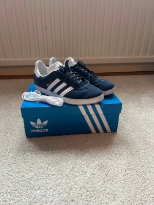 Nya Adidas Gazelle   - Snygga Adidas Gazelle sneakers i mörkblå mocka med vita detaljer och klassiska tre ränder på sidan. Endast använda en gång. nyskick. Stl 43