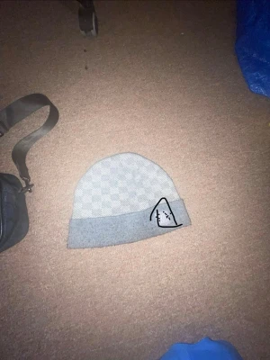 Ljusgrå mössa  - Säljer en ljusgrå mössa med diskret rutmönster och en bredare enfärgad kant. På kanten finns en liten patch med ett stiliserat bergmotiv. Perfekt för dig som gillar enkel men snygg stil.