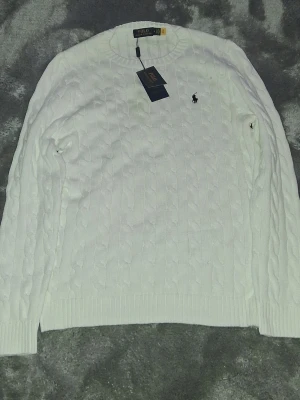 Vit kabelstickad tröja Polo Ralph Lauren - Säljer en vit kabelstickad tröja från Polo Ralph Lauren med klassisk rund halsringning och svart broderad logga på bröstet. Tröjan har ribbade muddar vid ärmslut och nederkant. Perfekt för dig som gillar stilrena och tidlösa plagg.