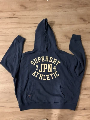 Sällsynt Blå Superdry hoodie med tryck - Mörkblå hoodie från Superdry med stor gul text på ryggen där det står 'Superdry JPN Athletic'. Klassisk känguruficka framtill och huva. Perfekt för en avslappnad och sportig stil. EXTREMT SÄLLSYNT MODEL SOM INTE SÄLJS LÄNGRE.