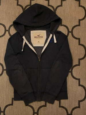 Blå zip-hoodie från Hollister - Blå hoodie från Hollister med dragkedja framtill, huva med vita snören och två fickor. Klassisk och enkel design med mjukt material. Perfekt för en avslappnad stil.