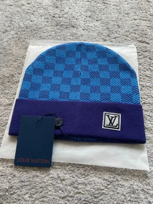 LVmössa - Snygg blå mössa med klassiskt rutmönster i två nyanser av blått. Uppvikt kant i mörkblått med logga framtill. Tillverkad i mjukt stickat material som känns lyxigt och trendigt. Perfekt accessoar för dig som vill sticka ut.