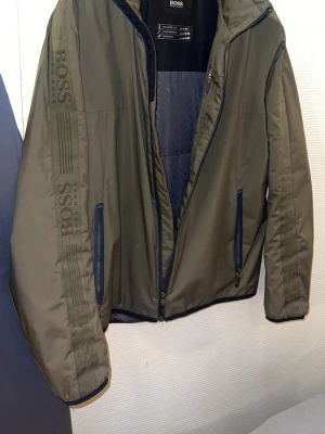 Olivgrön vårjacka från BOSS - Snygg olivgrön jacka från BOSS med diskret logga och ränder på ärmen.  hög krage och två fickor framtill. Perfekt för dig som vill ha en stilren och sportig look. Materialet är troligtvis polyester. Storlek M . Riktigt snygg jacka använd en gång. Hade behållit den om den inte vore för liten har för mig. Söker även likadan i samma färg eller annan om nån skulle vilja sälja också.  Pris kan diskuteras skriv för andra bilder