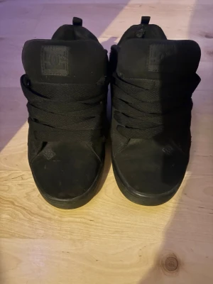 Svarta DC sneakers med hög plös - Säljer ett par helsvarta DC sneakers med hög plös och tjocka svarta skosnören. Skorna har en klassisk rund tå och är tillverkade i mocka med en platt sula. Perfekta för dig som gillar streetwear och skate-inspirerad stil. Med lite slitning på hälen