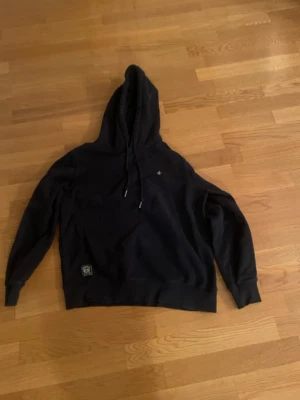 Svart hoodie från Morris - Snygg svart hoodie från Morris med klassisk huva . Hoodien har en liten logga på bröstet och en patch nertill vid midjan. Tillverkad i mjukt material som känns skönt mot huden. Perfekt för en avslappnad och stilren look. Är s men passar även xs 