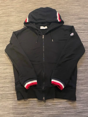 Svart Moncler hoodie med dragkedja - Svart hoodie från Moncler med hel dragkedja, huva och ribbade muddar med röd, vit och blå rand. Klassisk Moncler-logga på ärmen och snörning i huvan. Perfekt för en sportig och stilren look. Enda defekten är att lappen sitter lite löst och att en av metallbitarna på snörena saknas. Nfc-Scan funkar samt qr-koden!😁