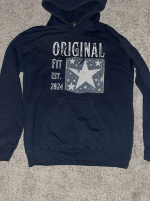 Mörkblå hoodie med stjärntryck - Säljer en mörkblå hoodie med stor vit stjärna och texten 'ORIGINAL FIT EST. 2024' på bröstet. Hoodien har huva med dragsko och en klassisk magficka. Perfekt för dig som gillar streetwear och vill ha en chill look.