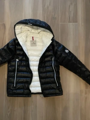 Svart dunjacka från Moncler med huva - Snygg svart dunjacka från Moncler med vit insida och huva. Jackan har två dragkedjeförsedda fickor framtill, quiltad design och Moncler-logga på ärmen. Perfekt för kalla dagar och riktigt stilren look.