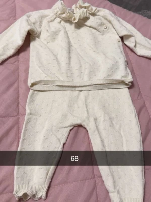 Vit stickad babyset med volangkrage - Sött tvådelat babyset i mjuk vit stickad bomull. Tröjan har lång ärm, volangkrage och små diskreta prickar i strukturen. Byxorna har resår i midjan och vågig kant vid benslut. Perfekt för mysiga dagar.