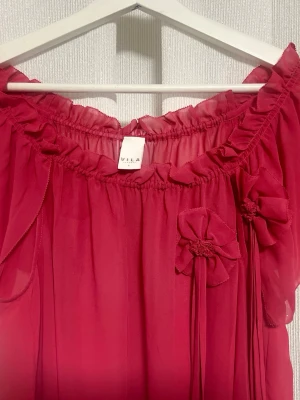 Röd volangklänning från Vila - Blus från VILA. Endast använd ett par gånger och i mycket gott skick. Säljer en röd blus från Vila med volangdetaljer och dekorativa blommor framtill. Klänningen har en lös passform, resår vid halsringningen och är gjord i ett tunt, transparent tyg med ett romantiskt uttryck. Perfekt för dig som gillar feminina detaljer.