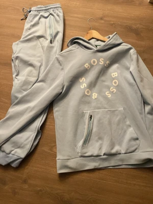 Hugo boss tracksuit - Säljer ett ljusgrått loungewear-set från BOSS med hoodie och matchande byxor. Hoodien har magficka med dragkedja och BOSS-tryck i cirkel på bröstet. Byxorna har resår i midjan och dragkedjeficka. Mjukt material, perfekt för chill dagar hemma.