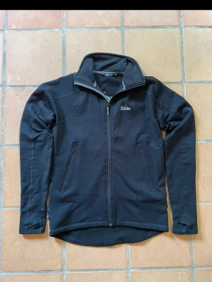 Svart fleecejacka från Tierra - Svart fleecejacka från Tierra med dragkedja framtill och två sidofickor med dragkedja. Jackan har hög krage och Tierra-logga broderad på bröstet. Perfekt för kyliga dagar och enkel att matcha med olika outfits.