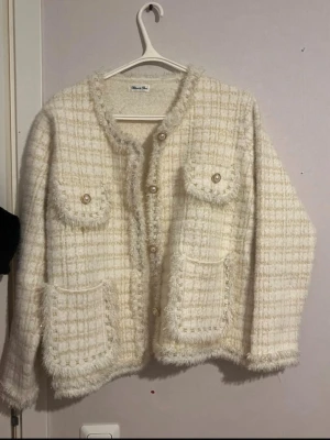 Vit rutig cardigan jacka Bleuet de Paris - Mysig vit cardigan jacka från Bleuet de Paris med rutigt mönster, dekorativa knappar och fyra fickor framtill. Jackan har lång ärm, fransiga kanter och en fluffig, stickad känsla som ger en lyxig vibe. knappt användt passar M och L