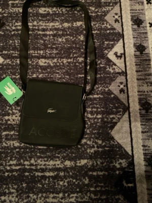 Svart axelväska från Lacoste - Snygg svart axelväska från Lacoste med präglad logga på framsidan och klassisk krokodil-detalj. Väskan har justerbar axelrem och stängs med magnetknappar. Perfekt för att bära det viktigaste med stil.