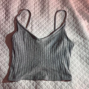 Grå ribbad croppad linnetopp - Säljer en grå croppad linnetopp med smala axelband och v-ringning. Toppen är ribbad och har en tight passform som sitter snyggt på kroppen. Perfekt till jeans eller kjol! :)