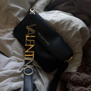 Svart axelväska från Valentino - Snygg svart axelväska från Valentino i skinn med grov struktur. Väskan har ett stort guldfärgat VALENTINO-spänne på axelremmen och diskret logga på framsidan. Rymligt med utrymme!Perfekt för dig som vill ha en statementväska med lyxig känsla!😊