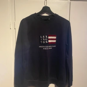 Mörkblå Lexington sweatshirt  - Snygg mörkblå sweatshirt från Lexington med broderad logga och amerikansk flaggdetalj på bröstet. Tröjan har rund hals och långa ärmar, perfekt för dig som gillar klassisk och stilren design. Passar dig som är ca 15 år.