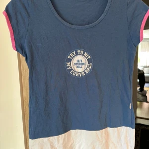 Blå t-shirt med tryck och rosa detaljer strl: M - Snygg blå t-shirt med rund hals och korta ärmar med rosa kant. Framtill finns ett broderat tryck med texten 'Try to hit my curve ball' och en baseboll. Nederdelen av t-shirten är vit, vilket ger en cool kontrast. Perfekt för dig som gillar sportiga och avslappnade plagg.