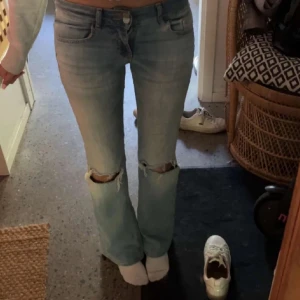 Ljusblå bootcut jeans med hål - Säljer ett par ljusblå jeans med bootcut-modell och slitna hål över knäna. Jeansen har klassisk femficksdesign, låg midja och är tillverkade i mjukt denimtyg. Perfekta för en avslappnad och trendig look.