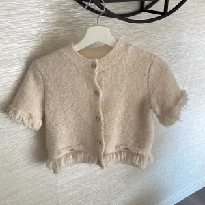 Beige kort kofta med eleganta knappar - Superfin beige kofta med kort passform och fluffiga detaljer vid ärmslut och nederkant. Koftan har glittriga pärlknappar framtill och på fickorna, samt korta ärmar. Materialet är mjukt och stickat, perfekt för att ge en mysig vibe. Storlek S men passar även XS