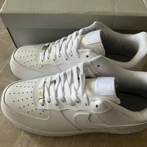 Nike Air Force 1 helvita sneakers - Klassiska Nike Air Force 1 sneakers i helvitt skinn med vit sula och vita skosnören. Ikonisk design med perforerad tå, metallbricka med 'AF1' och broderad Nike-logga på tungan. Perfekta för dig som gillar en clean och tidlös look.