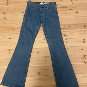 Blå bootcut jeans från H&M, stl 158 - Snygga blå jeans från H&M med bootcut och låg midja. Byxorna har justerbar midja och klassiska fickor både fram och bak. Tillverkade i bomull och polyester med lite stretch för extra komfort. Perfekta för dig som gillar retrovibbar och en avslappnad look.