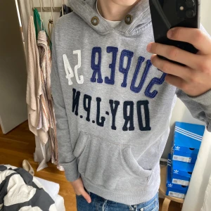 Grå hoodie från Superdry med tryck - Säljer en grå hoodie från Superdry med broderat tryck i blått, vitt och svart på bröstet. Tröjan har huva med snörningshål i metall, känguruficka och ribbade muddar. Perfekt för dig som gillar sportig och avslappnad stil. Tröjan säljs inte längre och går inte att få tag på!