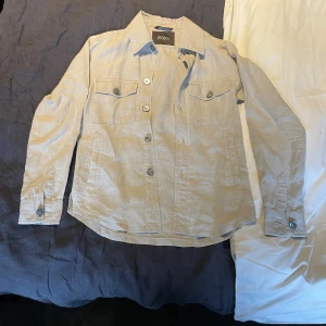 Beige overshirt från Oscar Jacobson  - Snygg beige overshirt från Oscar Jacobson i storlek XS. Modellen Maverick Jacket har klassisk krage, två bröstfickor med knapp och knäppning framtill. Tillverkad i linne, perfekt för dig som gillar stilrena och avslappnade plagg.