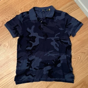 Camouflage piké från Polo Ralph Lauren - Snygg pikétröja från Polo Ralph Lauren i mörkblått och grått camouflage-mönster. Klassisk krage, korta ärmar och slim fit passform. Perfekt för dig som vill sticka ut med en cool streetstyle-look. Sitter som XS