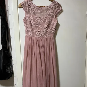 Rosa långklänning med spetsdetaljer - Supersöt långklänning i ljusrosa med vackert spetsliv och blommiga broderier upptill. Kjolen är mjukt fallande och i tunt tyg. Klänningen har ärmlös design och knappar i ryggen. Perfekt för speciella tillfällen!